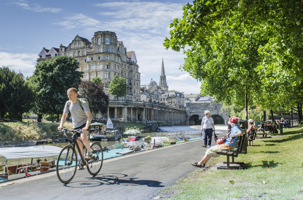 View of Bath, UK. Showing Pulteney Bridge and River Avon; The Weir; Man cycling alongside the River Avon with boats moored in background. Empire Hotel. Image copyright University of Bath.
